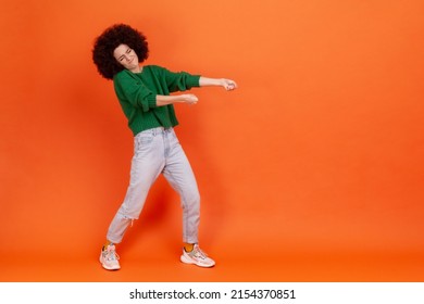 Full length profile portrait of woman with Afro hairstyle in green sweater pulling invisible heavy burden, striving hard to achieve success. Indoor studio shot isolated on orange background.