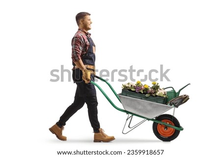 Full length profile shot of a gardener pushing a wheelbarrow with flowers and watering can isolated on white background