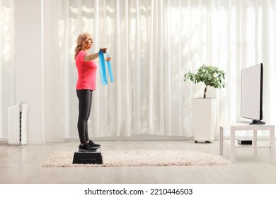 Full Length Profile Shot Of A Mature Woman Standing On An Aerobic Stepper And Exercising With An Elastic Band In Front Of Tv At Home