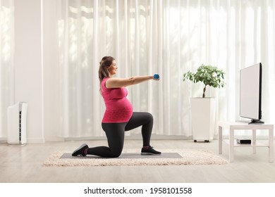 Full Length Profile Shot Of A Pregnant Woman Kneeling And Exercising With Dumbbells In Front Of Tv