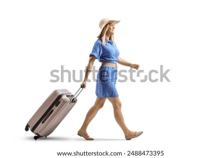 Full length profile shot of a woman in a blue dress and straw hat walking and pulling a suitcase isolated on white background
