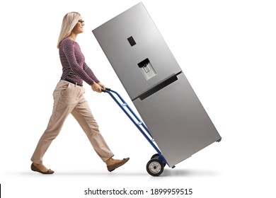 Full length profile shot of a young woman pushing a hand-truck with a new silver fridge isolated on white background