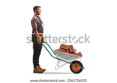 Full length profile shot of a young man standing with a wheelbarrow full of bricks isolated on white background
