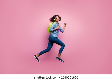 Full Length Profile Side Photo Of Cheerful Afro American Girl College Student Jump Run Hurry Lecture Listen Music Headset Wear Denim Jeans Shirt Bag Travel Isolated Pink Color Background