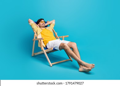 Full length profile side photo of positive guy traveler relax rest beach sun bathing lie comfort deckchair stretch hands wear white shorts isolated over blue color background