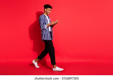 Full length profile side photo of young afro guy go walk happy chat type sms smartphone wear pants isolated over red color background - Powered by Shutterstock