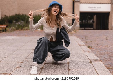 Full length radiant young caucasian girl kneels down against street. Blonde woman is wearing wide pants cargo, sweater top with buttons and blue panama hat, bag on shoulder. Girl look funky, fix hair.