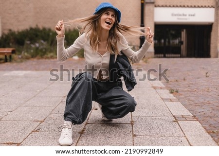 Full length radiant young caucasian girl kneels down against street. Blonde woman is wearing wide pants cargo, sweater top with buttons and blue panama hat, bag on shoulder. Girl look funky, fix hair.