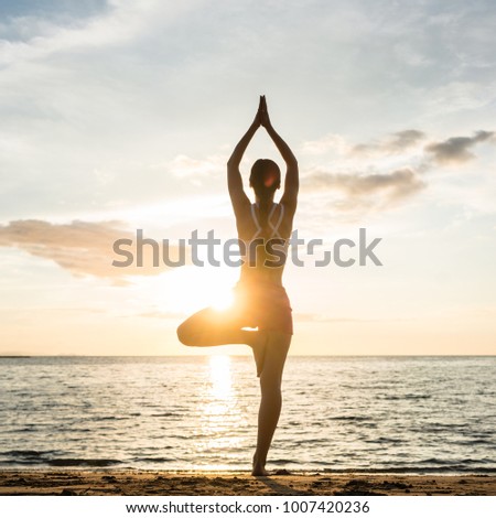 Full length rear view of the silhouette of a woman, standing on one leg while practicing the tree yoga pose on a tranquil beach at sunset during summer vacation in Indonesia