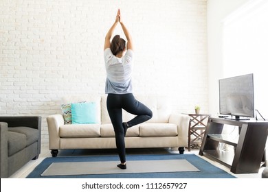 Full length rear view of young woman balancing on one leg, doing tree yoga pose and meditating in her living room
