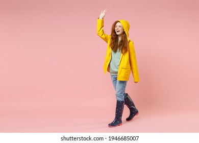 Full length redhead fun young woman in yellow waterproof hood raincoat outerwear walk go waving hand greet isolated on pastel pink background studio Outdoors lifestyle wet fall weather season concept