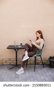 full length of redhead woman in turtleneck using smartphone near laptop on summer terrace