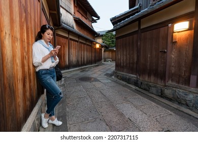 full length of relaxed asian Japanese girl visitor leaning against wooden wall and looking at her cellphone in illuminated Ishibeikoji alley in gion Kyoto japan at sunset - Powered by Shutterstock