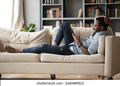 Full Length Relaxed Young African Ethnicity Man In Glasses Lying On Comfortable Couch, Enjoying Spending Weekend Leisure Time Chatting In Social Network On Phone With Friends, Watching Funny Videos.