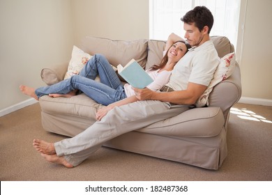 Full Length Of A Relaxed Young Couple Reading Book On Couch At Home