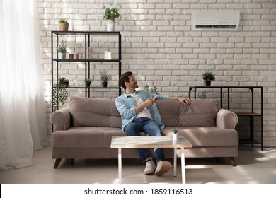 Full Length Relaxed Young Man Sitting On Comfortable Sofa, Turning On Climate Control System With Remote Controller, Feeling Satisfied With Fresh Conditioned Air During Hot Summer Time Weather At Home