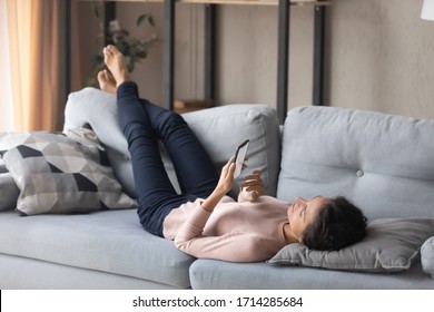 Full length relaxed young woman lying on comfortable couch on soft pillow, using mobile applications. Happy lady chatting in social network with friends, enjoying spending leisure time at home. - Powered by Shutterstock