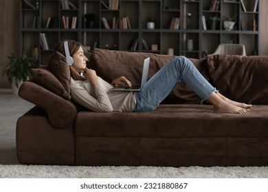 Full length relaxed young woman wearing wireless headphones, watching funny movies on laptop or listening music, lying barefoot on cozy sofa, enjoying stress free leisure weekend time at home.
