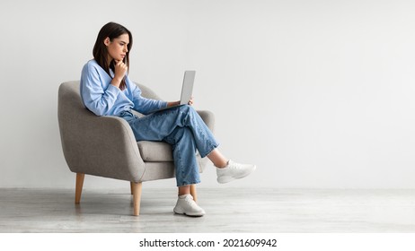 Full length of serious young woman looking at laptop screen, sitting in armchair, having problem with online work or studies against white studio wall, banner with copy space