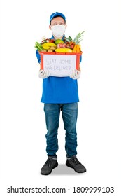 Full Length Of Shipper Wearing Face Mask While Holding A Box Full Of Groceries With Text Of Stay Home We Deliver And Standing In The Studio. Isolated On White Background