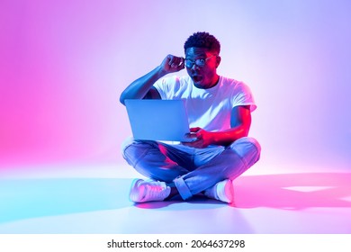 Full length of shocked black guy sitting cross legged with laptop pc, touching his glasses, winning online lottery in neon light. Web casino, remote job promotion, huge internet store sale