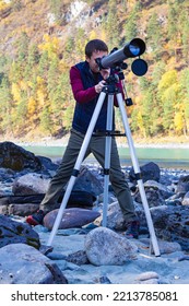 Full Length Shoit An Adult Attractive Man In Sportswear Looking Through Telescope On Bank Of Mountain River.Outdoor Astronomy, Stargazing, Selective Focus, Copy Space