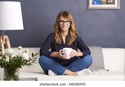 Full Length Shot Of An Attractive Middle Aged Woman Holding Hand A Mug With Tea While Relaxing On Sofa At Home.