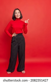 Full Length Shot Of Beautiful Asian Woman In Turtleneck And Black Trousers Stands Bare Feet Indicates Away On Blank Copy Space Shows Advertisement Or Promo Attracts Your Attention To Something