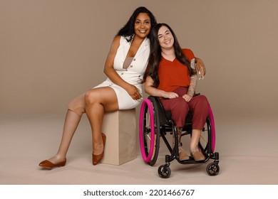 Full length shot of a Black woman in her 20's with an amputated leg and a Caucasian woman in her 20's - wheelchair user smiling together on a neutral background. Stock Photo