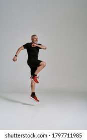 Full Length Shot Of Cheerful Middle Aged Muscular Man In Black Sportswear Jumping While Having Workout In Studio Over Grey Background. Sport, Healthy Lifestyle Concept. Vertical Shot