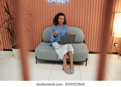 Full length shot of cheerful young multiethnic woman presenting during online conference joining video call via laptop, while sitting on comfy couch in lounge area of modern coworking space - Powered by Shutterstock