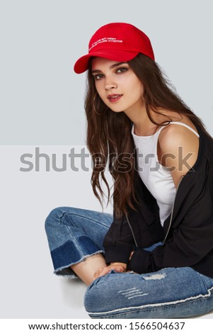 Full length shot of a dark-haired girl, sitting on a floor and wearing red baseball cap with lettering 