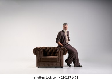 Full length shot of fashionable mature man with beard wearing elegant suit looking at camera, leaning on armchair while posing isolated over light background