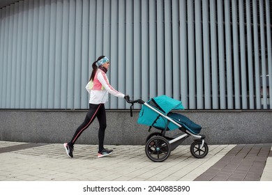 Full length shot of female runner mom training with a stroller in the city on a cold autumn day. Parenting, healthy lifestyle, sports concept. Side view