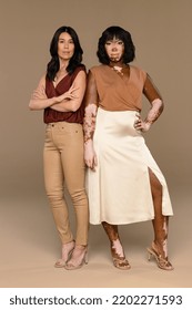 Full Length Shot Of A Filipino Woman In Her 50's And An African American Woman With Vitiligo In Her 30's Standing Confidently On A Neutral Background.