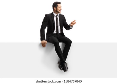 Full Length Shot Of A Handsome Young Man In A Suit Sitting On A Blank Panel And Gesturing With Hand Isolated On White Background