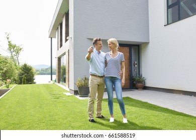 Full Length Shot Of A Happy House Owners Standing In The Garden And Holding A Key After Moving Their New Home. 