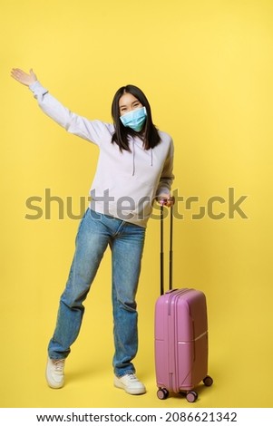 Full length shot of happy korean girl tourist on vacation, posing with suitcase in medical face mask, enjoying travelling, concept of tourism and pandemic healthcare