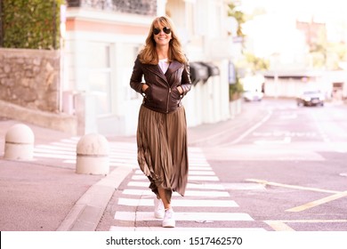 Full Length Shot Of Happy Middle Aged Woman Walking On The Street While Looking At Camera And Smiling. Confident Woman Wearing Sunglasses And Biker Jacket With Skirt. 