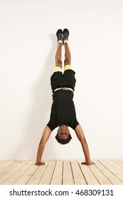 Full length shot of a happy smiling attractive young black model in black and yellow workout clothes doing handstand against a white wall on wooden floor.