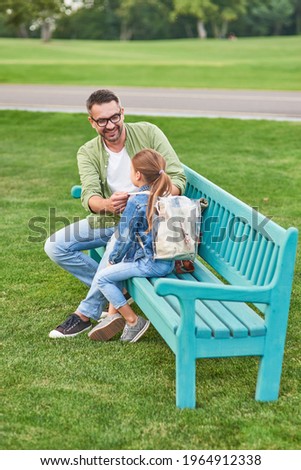 Full length shot of happy young father and his little daughter sitting on the bench, spending time together in the green park on a warm day