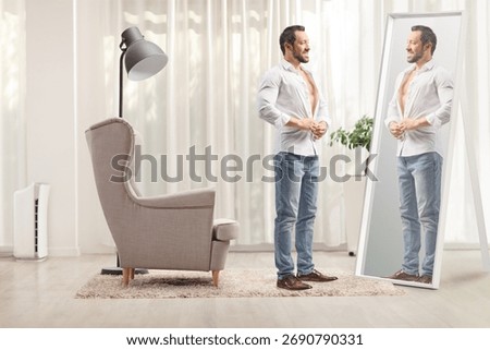 Full length shot of a man buttoning a white shirt in front of a mirror in a room
