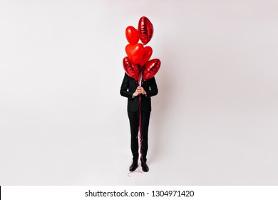 Full length shot of man hiding behind air balloons. Studio photo of guy in suit posing in valentine's day.