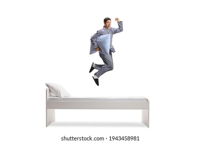 Full length shot of a man in pajamas holding a pillow and jumping on a bed isolated on white background