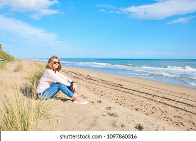 Full Length Shot Of Middle Aged Woman (wearing Jeans, Leather Jacket And Sunglasses) Sitting  At The Beach And Relaxing.