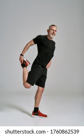 Full Length Shot Of Middle Aged Muscular Man In Black Sportswear Looking At Camera While Stretching Body Before Workout In Studio Over Grey Background. Sport, Healthy Lifestyle Concept. Vertical Shot