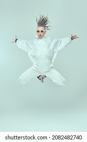 Full length shot of a modern punk rock musician with dreadlocks in white overalls and stylish glasses  jumping on a light background with his arms streche aside. Youth alternative culture. 
