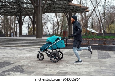 Full Length Shot Of Motivated Sportive Dad In Sportswear Running With A Pram Along An Empty Park On A Cold Autumn Day. Parenting, Healthy Lifestyle, Sports Concept. Side View