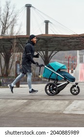 Full Length Shot Of Motivated Sporty Dad Running And Carrying A Stroller With A Child While Training In The Park On A Cold Autumn Day. Parenting, Healthy Lifestyle, Sports Concept. Side View