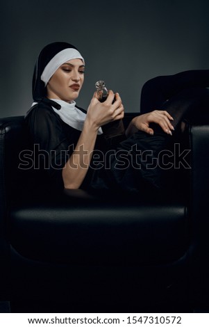 Full length shot of a nun, sitting on a chair. She's holding bottle of wine in her hand and looking with disgust on it and wrinkles her nose. She's sitting with her legs crossed. 